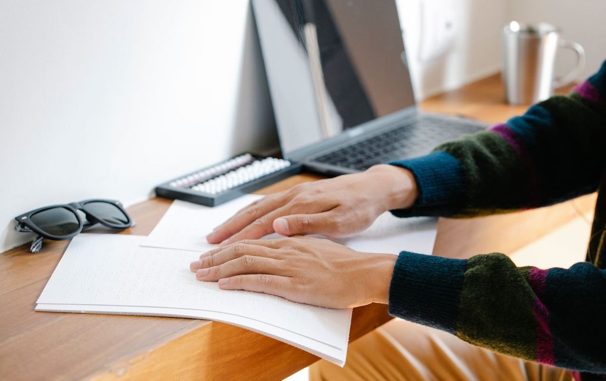 Deux mains posées sur une page imprimée en braille, devant un clavier d'ordinateur